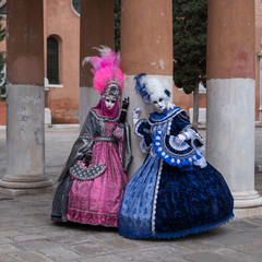 Fototapeta na wymiar Two women in masks and ornate blue and pink costumes standing in front of pillars at a monastery during Venice Carnival (Carnival di Venezia).
