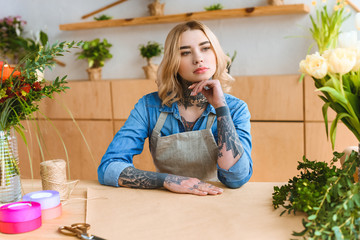 pensive young florist in apron sitting at workplace and looking away in flower shop