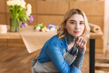 young tattooed woman in apron sitting with hand on chin and looking at camera