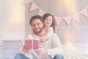 Looking forward to celebration. Attentive handsome enthusiastic man sitting on a couch waiting for his wife while his daughter hugging him from behind