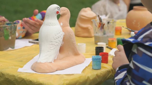 Unknown little boy sitting at table in park painting brushes on clay figure in form of dove. Development of art and painting in children. Clay pottery workshop. Close-up