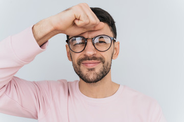Obraz premium Portrait of serious handsome young male with beard and mustache, keeps hand on forhead, wears pink clothes, isolated over white studio background. Hispter guy has some problems with work or projects