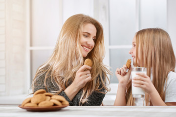 Obraz na płótnie Canvas Photo of two laughing sisters - younger and oder. Both girls have blonde straight hair. They are eating delicious sweet cookies from plate and drinking milk near white kitchen's table.