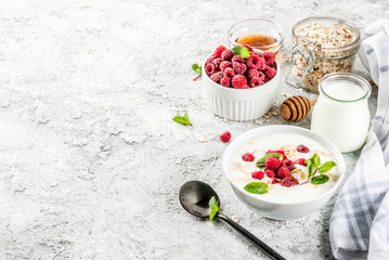 Raw ingredients for summer healthy breakfast, cereals (oats), fresh Raspberry, mint leaves, yoghurt, honey, on light concrete background, copy space
