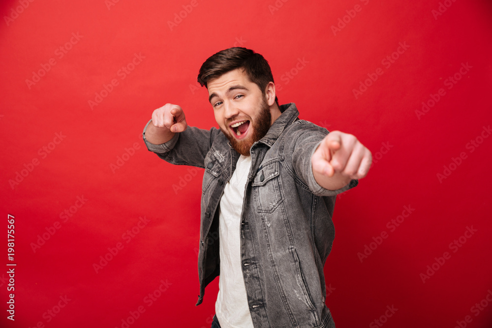 Canvas Prints Joyful bearded man 30s in jeans jacket looking on camera with pointing index fingers on you, isolated over red background