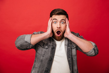 Photo of surprised man 30s wearing beard in jeans jacket grabbing head and reacting emotionally, isolated over red background