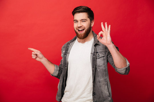 Photo of content brunette man 30s in jeans jacket showing ok sign and finger aside on copyspace, isolated over red background