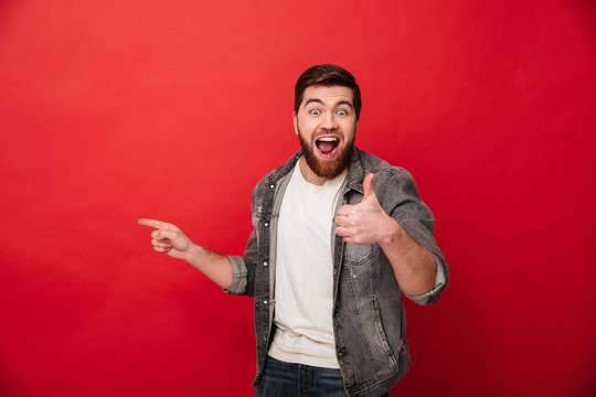 Delighted brunette man in casual clothin showing thumb up while demonstrating copyspace with finger aside, isolated over red background