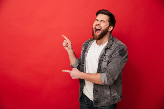 Photo of joyous man in casual clothing bursting in laughter and gesturing fingers on copyspace, isolated over red background