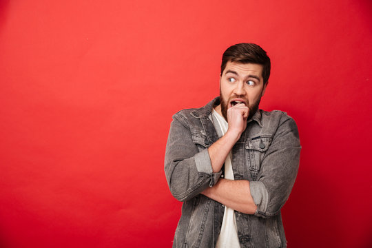 Image of scared man 30s in jeans jacket looking aside on copyspace and biting his fist in horror or fright, isolated over red background