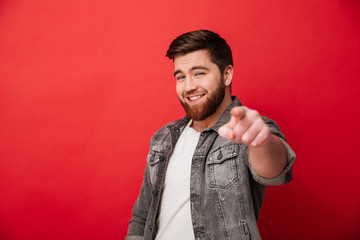 Photo of handsome cheerful man 30s in jeans jacket smiling and pointing index finger on camera,...