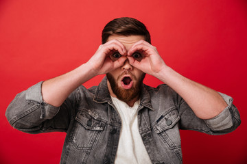 Portrait of joyful guy 30s in jeans jacket having fun and looking on camera through holes like binoculars, isolated over red background