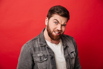 Portrait of amusing guy 30s in jeans jacket winking and looking on camera with funny gaze, isolated over red background