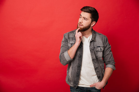 Portrait of serious man 30s in jeans jacket looking aside with brooding gaze on copyspace, isolated over red background