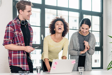 Three young colleagues laughing while standing in front of a laptop in the meeting room of a modern...
