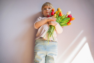 Powazna little girl o blond wlosach trzyma bukiet kolorowych tulipanow w rekach w slonecznym pokoju w domu. Concept swieta. © AnastazjaSoroka