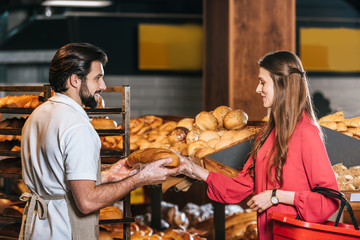 side view of shop assistant giving loaf of bread to woman with shopping basket in supermarket