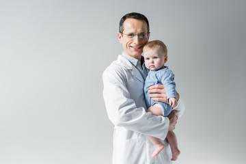 handsome adult pediatrician holding little baby and looking at camera isolated on grey