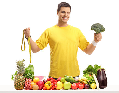 Young man holding a measuring tape and broccoli behind a table filled with fruit and vegetables