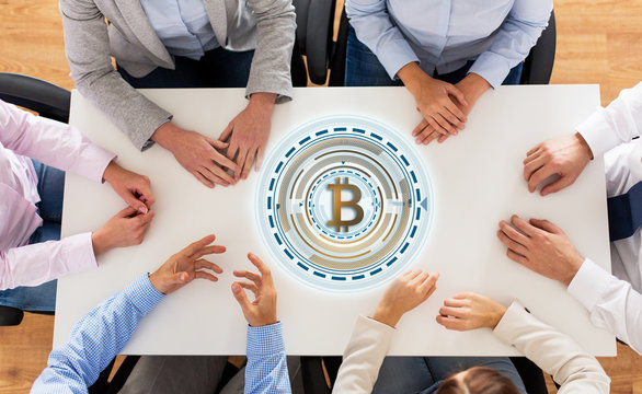 business, cryptocurrency and finance concept -team sitting at wooden table with bitcoin hologram in office, shot from top