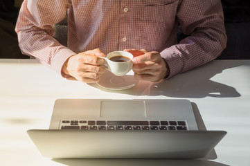 Coffee break in bright office. Male hands hold cup of steaming hot coffee next to laptop computer in minimalistic indoor room on sunny day