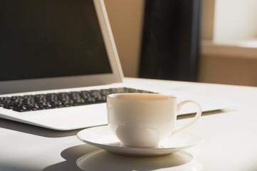 Cup of hot steaming morning coffee and laptop computer. Close-up view of PC and white espresso cup on white sun lit table at modern office or room