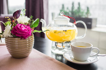 Glass teapot with hot pineapple tea with pineapple slices in a cafe. A cup of fragrant yellow tea, a glass teapot and beautiful pink flowers in a vase.