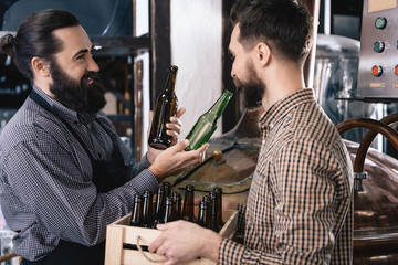 Bearded brewer picks up color glass beer bottle in brewery. Process of beer manufacturing.