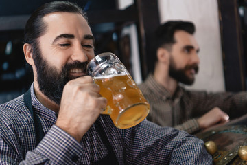 Bearded brewer in apron tastes light beer from beer mug at craft brewery. Examining taste of beer.