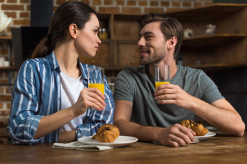 Smiling man and woman drinking juice and eating croissants by kitchen table
