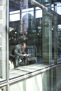Black business man texting while sitting in front of a large widow in a lobby waiting area.