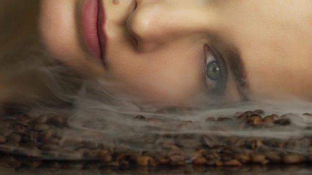 Beautiful young sexy woman portrait bright makeup decorative cosmetics face care lies on roasted coffee beans aromatic scent wraps her face with smoke and the taste open her eyes.