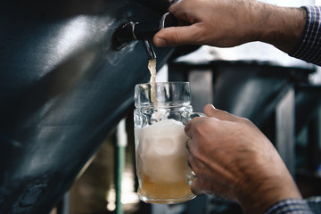 Strong man's hands pour beer in tumbler from beer tap at craft brewery.