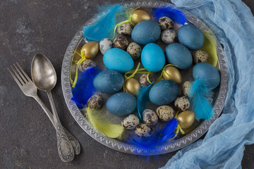 Easter composition: in a silver plate lay blue eggs, golden eggs, quail eggs and feathers, and cutlery - fork and spoon
