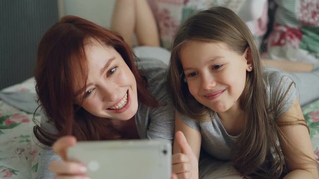 Happy mother and little girl taking selfie photo with smartphone camera and have fun grimacing while sitting in cozy bed at home