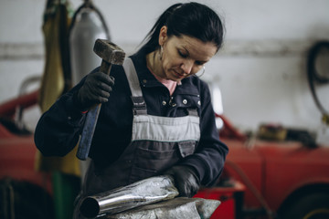 Strong and worthy woman doing hard job in car and motorcycle repair shop. She using hammer to fix broken exhaust.
