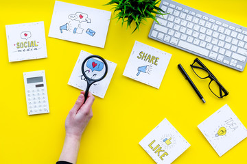 Social media promotion. Work desk with socail media icons. Yellow background top view. Hand with magnifier