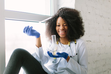 Young beautiful African American girl doctor in a white coat with a stethoscope. sitting at a table with reagent flasks on white background. Researcher researching in the laboratory