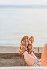 Close up of young woman resting her legs in leather sandals at sea shore, back view with arm and legs of unidentifiable woman in colorful skirt