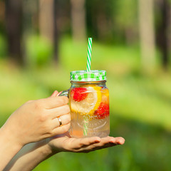 Young girl holding a jar of lemonade out of lemons and strawberries, with green lid and straw. A refreshing drink on a Sunny day.