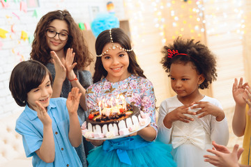 Little pretty girl with pendant chain on her head holds cake with candles at celebration of birthday.