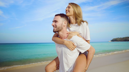 Beautiful happy couple having fun, doing piggyback on tropical sandy beach over sea and sky background