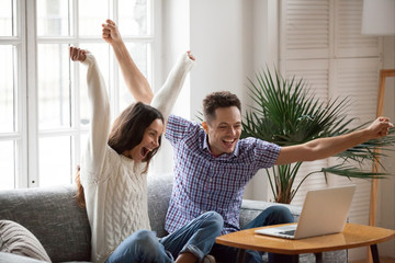 Excited man and woman screaming with joy raising hands looking at laptop screen sitting on sofa at...