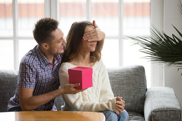 Loving husband or boyfriend closing eyes of wife girlfriend with hand presenting romantic surprise or birthday gift in pink box, family couple celebrating Valentines day anniversary together at home