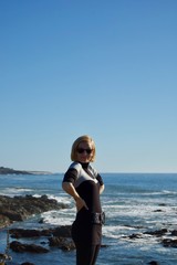 Happy blonde female model enjoys the beautiful scenic coastal view in California (USA): Untouched nature of the pacific ocean with limestone rock cliffs and crashing waves with a clear blue spring sky