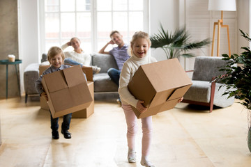 Fototapeta na wymiar Excited kids laughing playing in new home carrying boxes, happy family with children enjoying relocation, small girl and boy having fun in living room helping parents to pack, moving day concept