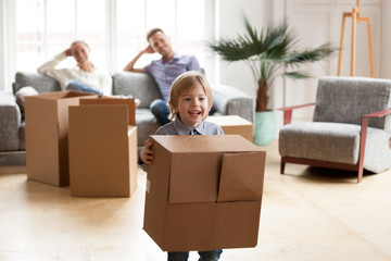 Fototapeta na wymiar Excited little boy playing with cardboard box on moving day, parents relaxing on sofa while their happy active son enjoying packing relocating into new home, cute kid having fun in modern living room