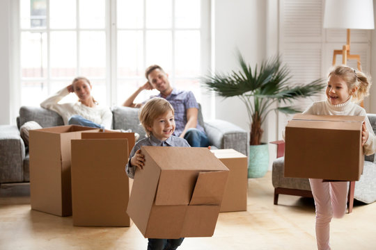 Cute small kids carrying boxes playing together on moving day concept, active happy boy and girl chasing each other in living room, excited children having fun helping parents pack unpack in new home