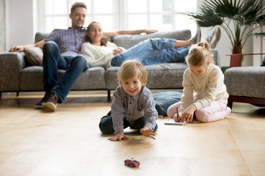 Cute kids playing while parents relaxing sofa at home together, smiling active boy entertaining with toy car near his sister on floor, happy family spending time together in living room on weekend