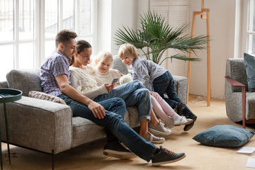 Happy family with children using mobile apps together at home, young couple and kids having fun playing game on smartphone sitting on sofa, parents and son daughter relaxing in living room with phone